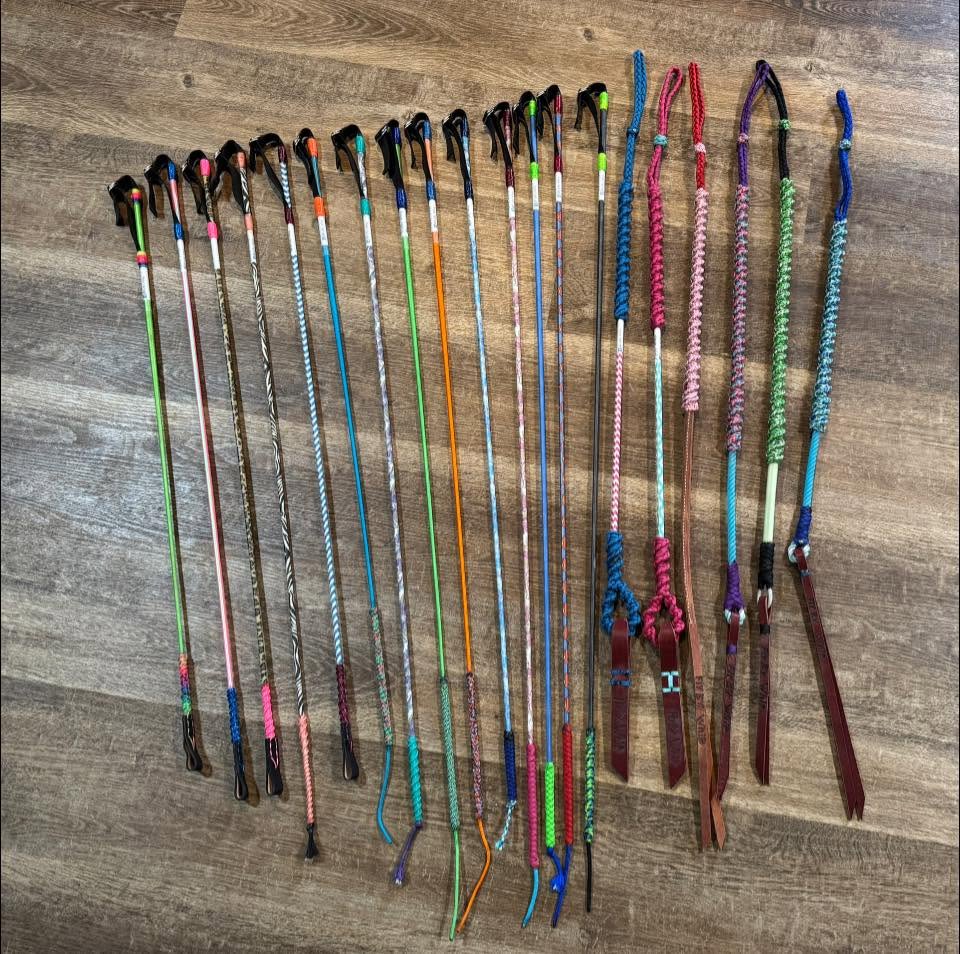 FlyBye Whips barrel racing whips in assorted colors arranged on a wood floor.