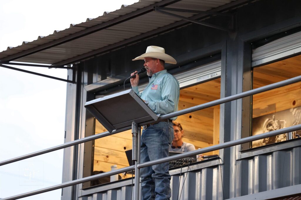 JR McLelland announcing from an elevated announcer&rsquo;s stand, speaking into a microphone at the railing.