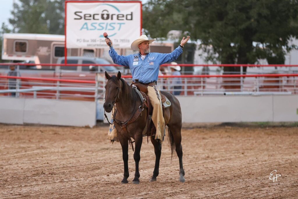 JR McLelland on a dark horse in a rodeo arena with his arms spread wide, a microphone in one hand.